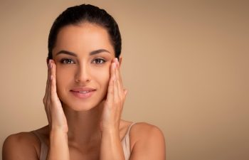 Antiage skincare. Beautiful millennial woman touching skin near eyes and looking at camera, lady with flawless skin standing over beige studio background, closeup portrait, panorama, copy space