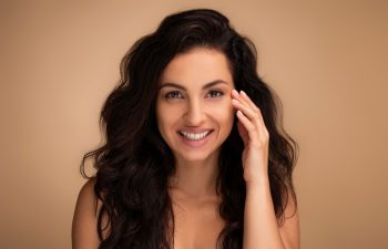 Anti-aging eye care treatment, aesthetic procedures concept. Attractive brunette long-haired young woman touching under eyes skin and smiling, closeup, beige studio background